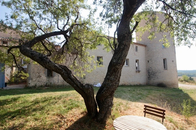 "Vue d'un arbre centenaire et d'une partie du Château de la Gabelle en Drôme provençale, avec une table et une chaise en bois pour se détendre dans le jardin luxuriant.